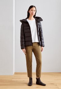 Chaqueta de plumas negra con capucha, diseño acolchado, llevada sobre una camiseta blanca. Pantalones beige y botines negros completan el conjunto.