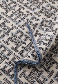 Geweven textiel met een geometrisch patroon in zwart en blauw op een licht crème achtergrond, met een blauwe gestikte rand en een zachte textuur.