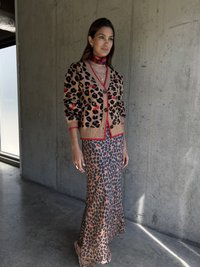 Beżowy kardigan dziergany w czarne i czerwone cętki lamparta, z zapięciem na guziki, na dopasowanej sukience w cętki lamparta. Modelka nosi czerwoną chustę i biżuterię.