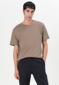 Bruine T-shirt met korte mouwen en een ronde halslijn, gemaakt van zacht, glad materiaal. Draagt het met donkere broek, handen in de zakken.