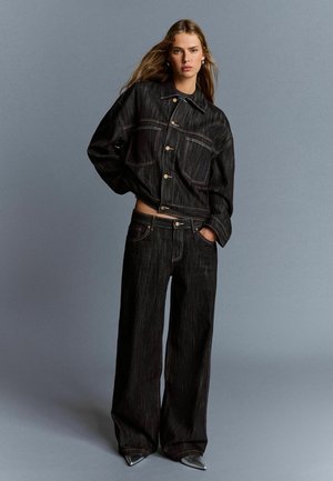 Woman with loose long hair wearing oversized black denim jacket and wide-leg jeans, posing against gray background.