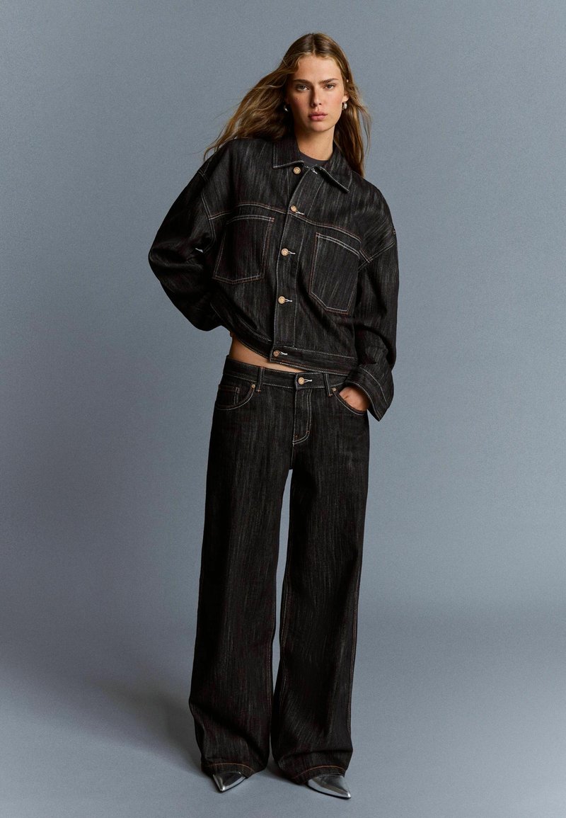 Frau mit langen, lockeren Haaren, die eine oversized schwarze Jeansjacke und weit geschnittene Jeans trägt, posiert vor einem grauen Hintergrund.