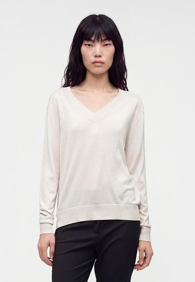 Femme aux longs cheveux noirs portant un pull beige clair à col en V et un pantalon noir, debout devant un fond blanc uni.