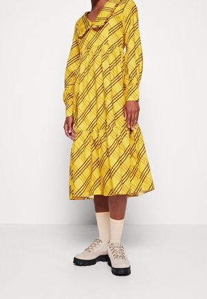 Kvinde iført en gul midikjole med brune diagonale striber, flæset krave, lange ærmer, beige sokker og chunky beige snøresko.