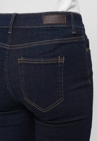 Jeans de mezclilla azul oscuro con un bolsillo trasero y etiqueta de parche de cuero. Presenta costuras amarillas y un diseño ajustado para un aspecto aerodinámico.