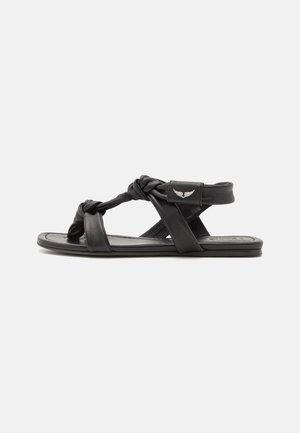 Sort flad sandal med snoede stropper og ankelrem med en lille metal detalje formet som en vinge på hvid baggrund.