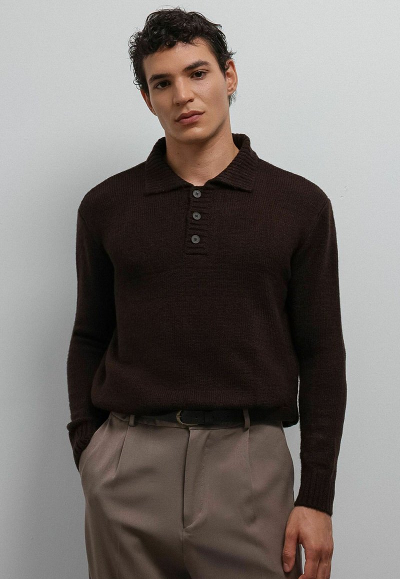 Jonge man met krullend haar, gekleed in een donkerbruine gebreide trui met lange mouwen en knopen, en beige geplooide broek met een zwarte riem.