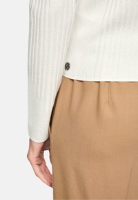 Heller, gerippter Pullover in Off-White mit abgerundetem Saum und einem dekorativen silbernen Knopf. Kombiniert mit beigen, plissierten Hosen.