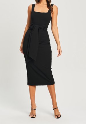 Frau modelliert ein schwarzes ärmelloses Midikleid mit quadratischem Ausschnitt und großer Taillenbinde, trägt schwarze, hochhackige Sandalen mit Riemchen.