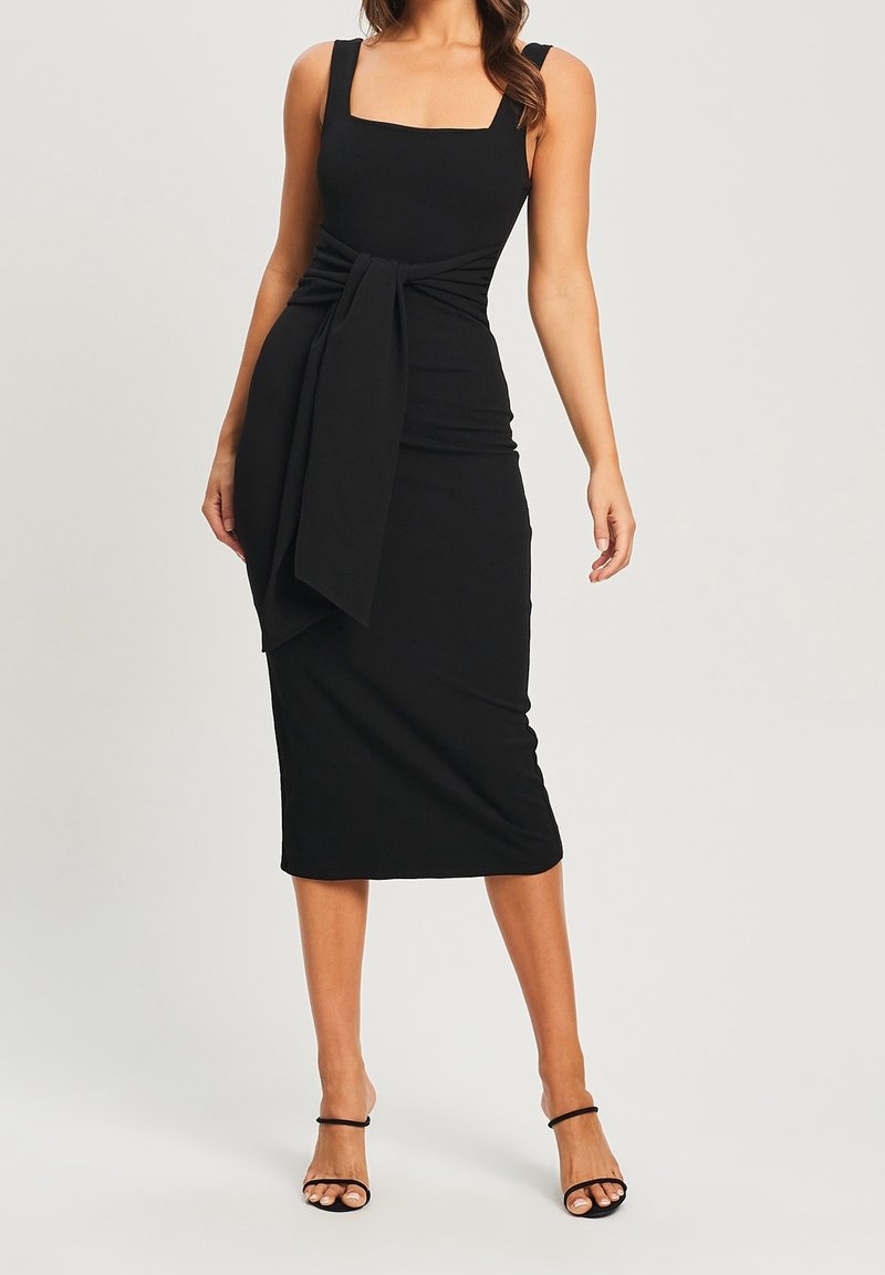 Femme portant une robe midi noire sans manches avec un décolleté carré et une large ceinture à la taille, portant des sandales noires à talons hauts à brides.