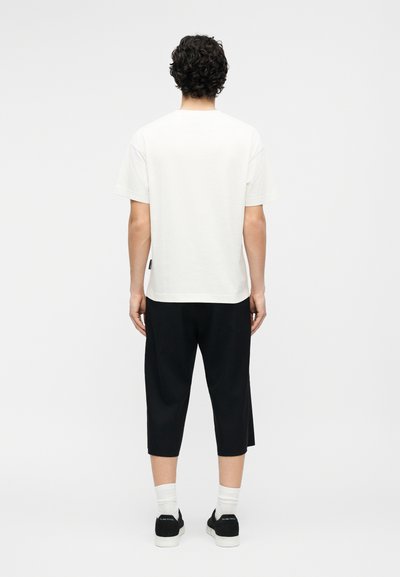 Άτομο με σγουρά σκουρόχρωμα μαλλιά που κοιτάζει προς τα πίσω, φορώντας λευκό κοντομάνικο πουκάμισο, μαύρο παντελόνι μέχρι τον αστράγαλο, λευκές κάλτσες και μαύρα αθλητικά παπούτσια.