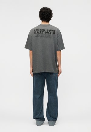 Oseba z kodrastimi lasmi, oblečena v ohlapno sivo majico s črnim napisom in modrimi kavbojkami, stoji obrnjena stran proti enobarvnemu ozadju.