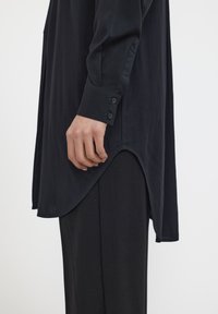 Chemise noire à manches longues avec des poignets boutonnés, un ourlet incurvé et une texture lisse, assortie à un pantalon noir.