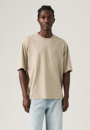 Mand iført en beige, oversized T-shirt med korte ærmer og et diskret emblem på brystet samt lyseblå jeans, stående foran en ensfarvet baggrund.