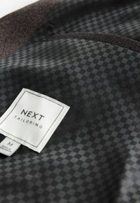 Black tailored jacket interior featuring a patterned fabric with a label reading "NEXT TAILORING," size M. Smooth texture, subtle check design.