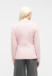 Γυναίκα με μακριά ξανθά μαλλιά, φορώντας ένα εφαρμοστό ανοιχτό ροζ σακάκι και φαρδιά σκούρα γκρι παντελόνια, με την πλάτη προς τα εμπρός, σε μονόχρωμο φόντο.