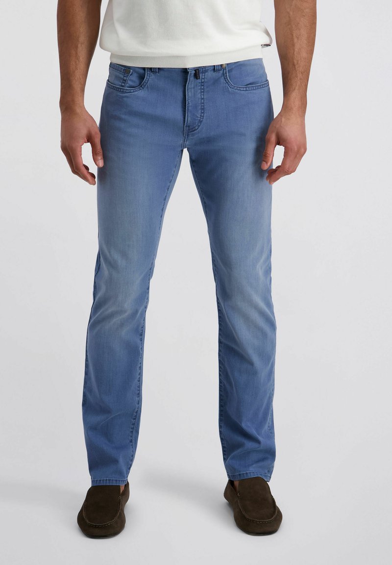 Mann trägt hellblaue Slim-Fit-Jeans, weißes Hemd und dunkelbraune Slipper, steht vor einem schlichten weißen Hintergrund.