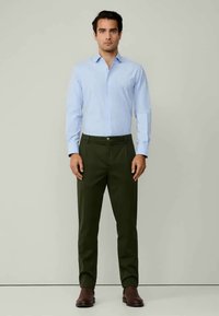 Ανοιχτό μπλε πουκάμισο με κουμπιά, πράσινα παντελόνια, καφέ δερμάτινα παπούτσια. Το πουκάμισο διαθέτει κολάρο, ενώ τα παντελόνια έχουν ίσια γραμμή.