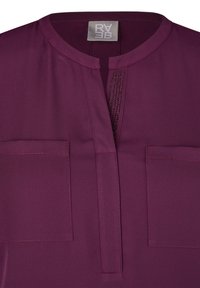 Pflaumenfarbene Bluse mit rundem Ausschnitt, halber Knopfleiste und zwei Fronttaschen. Mit subtilen Nähten und glatter Textur.