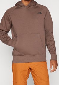 Felpa con cappuccio marrone, dotata di una tasca frontale, realizzata in tessuto morbido. Presenta maniche raglan e un piccolo logo nero sul petto.