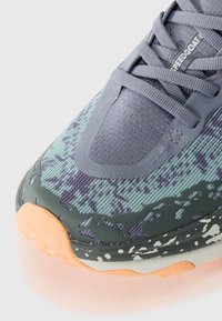 Chaussure de trail running avec un motif multicolore comprenant des nuances de vert, de gris et de violet, un dessus texturé et une semelle extérieure en caoutchouc orange.