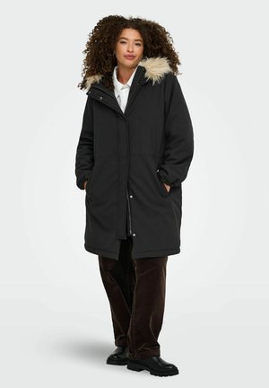 Femme aux cheveux bouclés portant un manteau d’hiver noir avec une capuche en fourrure, une chemise blanche, un pantalon foncé et des bottes noires, debout les mains dans les poches.