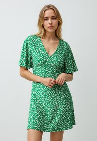 Groene bloemenjurk met een V-hals, korte fladdermouwen en een getailleerde corsage, met een herhalend wit bladerenpatroon op zachte stof.