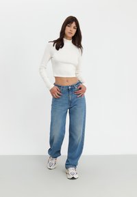 Hvit ribbet crop-top kombinert med lyseblå brede jeans og flerfargede treningssko, satt mot en nøytral bakgrunn.