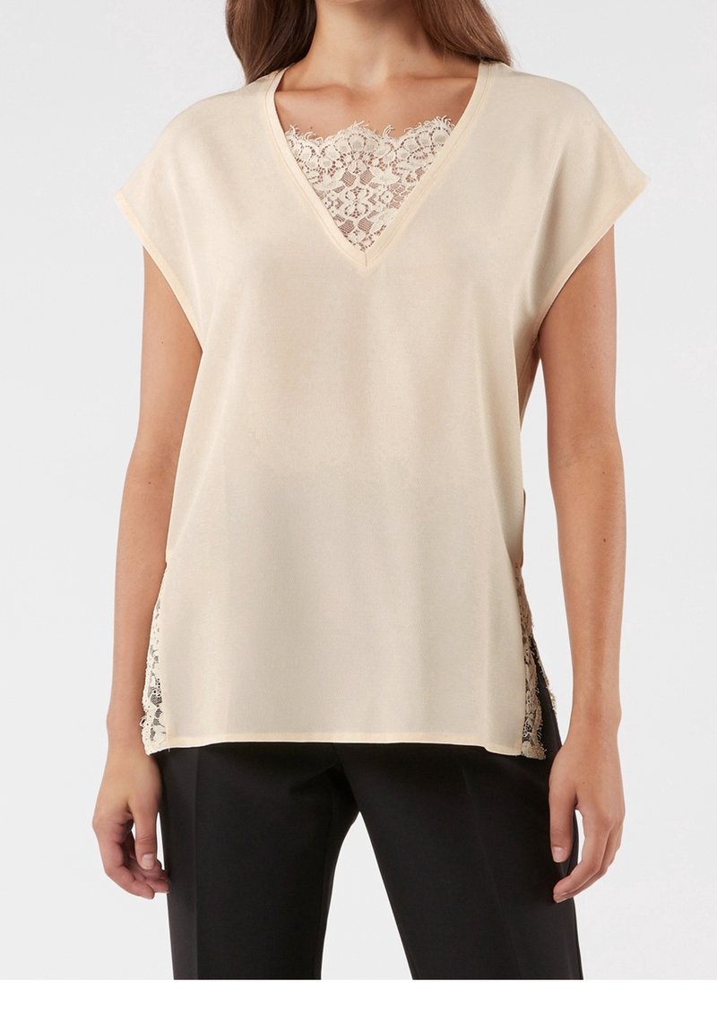 Blouse beige avec un col en V, ornée de détails en dentelle au plastron et sur les côtés. Texture douce, manches courtes et coupe décontractée.
