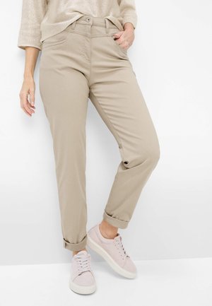 Person iført beige bukser med opsmøg, beige sweater og lys rosa sneakers stående mod en hvid baggrund.