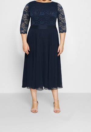 Woman wearing a navy blue midi dress with lace bodice and sleeves, a satin waist belt, and nude ankle-strap heels, standing against a light background.