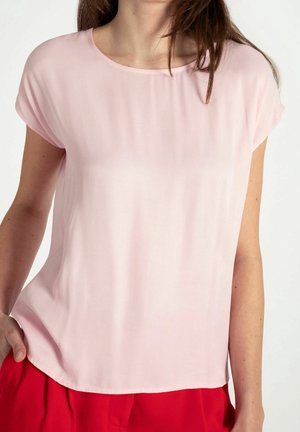 Frau trägt eine hellrosa kurzärmelige Bluse und rote Hose, mit der linken Hand auf der Hüfte, vor neutralem Hintergrund.