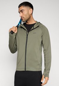 Λαδι πράσινο μπουφάν με φερμουάρ και κουκούλα, κατασκευασμένο από λείο ύφασμα. Διαθέτει μαύρες λεπτομέρειες και λογότυπο στο στήθος. Το μοντέλο κρατά ρακέτα τένις.