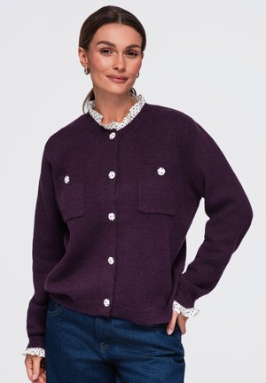 Femme aux cheveux bruns portant un cardigan violet boutonné par-dessus un chemisier blanc à pois et un jean bleu, debout avec une main sur la hanche.
