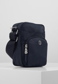 Kompaktowa granatowa torba na ramię z regulowanym paskiem, przednią kieszenią na zamek, srebnym uchwytem zamka błyskawicznego oraz małym, okrągłym emblematem logo.