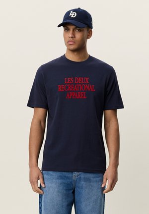 Jeune homme portant une casquette de baseball bleu marine et un t-shirt bleu marine avec un texte rouge "LES DEUX RECREATIONAL APPAREL" et un jean bleu, debout devant un fond uni.