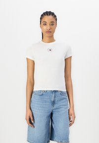 Camiseta de algodón blanca con mangas cortas, que presenta un pequeño logo rojo y azul en el pecho. Combinada con pantalones cortos de mezclilla azul claro.