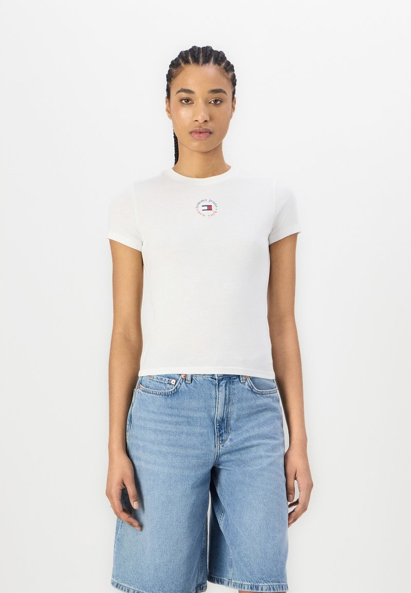 White cotton t-shirt with short sleeves, featuring a small red and blue logo on the chest. Paired with light blue denim shorts.