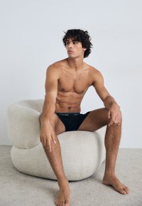 Boxeri scurți negri din bumbac, cu un elastic pe care este inscripționat logo-ul "Calvin Klein", purtați de un model masculin așezat pe un fotoliu texturat de culoare crem.
