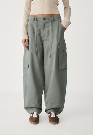 Cargo pants in light green, featuring two large side pockets, drawstring waist, and a loose fit. Paired with brown sneakers and a cream top.