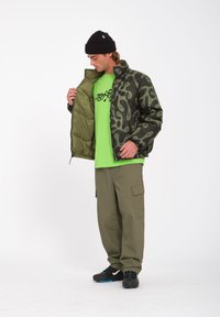 Groene gewatteerde jas met een rits, gedragen over een felgroen T-shirt, gecombineerd met losse olijfgroene cargobroek en zwarte sneakers.
