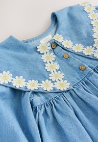 Robe en denim avec un tissu bleu clair, ornée d'un motif de broderie florale avec des marguerites blanches au centre jaune, et des boutons en bois.