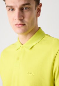 Polo tričko v jasne žltej farbe s textúrovanou tkaninou, klasickým golierom, gombíkovou légou a vyrazeným logom "BOSS" na hrudi.
