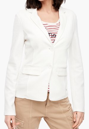 Žena oblečená v bílém texturovaném blejzru přes červeno-bíle pruhovanou košili v kombinaci s béžovými kalhotami, zobrazená od poloviny trupu po stehna.