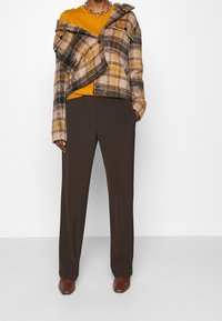 Brązowa kraciasta, oversize'owa kurtka z guzikami z przodu, noszona na musztardowej koszuli. Brązowe spodnie prostego kroju i brązowe botki dopełniają stylizację.