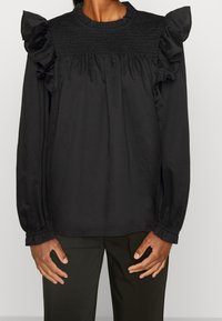 Blusa nera a maniche lunghe con scollatura e maniche arricciate, caratterizzata da dettagli arricciati sul corpetto. Tessuto morbido e leggero.