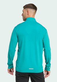 Tyrkysový sportovní top s dlouhým rukávem a vysokým límcem, texturovaná látka a minimalistický stříbrný akcent na spodním lemu, nošený osobou zezadu.