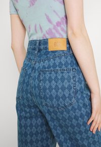 Niebieskie dżinsy z denimu z diamentowym wzorem, z wysokim stanem i brązową skórzaną naszywką z logo z tyłu. Widoczne dwie tylne kieszenie.