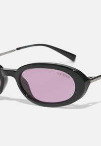 Svarta solglasögon med ovala bågar, rosa linser och silvermetalldetaljer som har "GUESS"-logotypen på en av linserna.