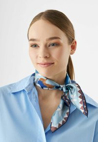 Foulard en soie avec un fond bleu et des motifs floraux et géométriques en marron et blanc. Attaché autour du cou, avec une texture douce.